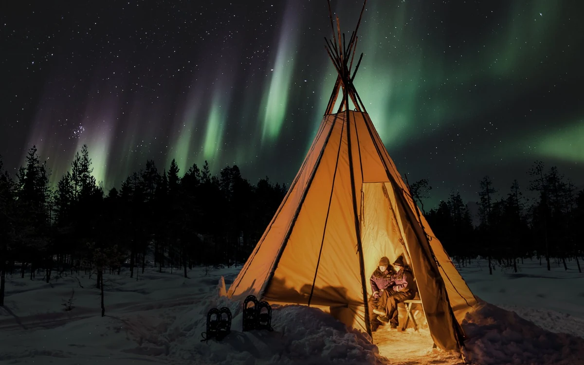 A lit canvas lavvu tent with two people sitting inside and snowshoes in the snow, under a green sky