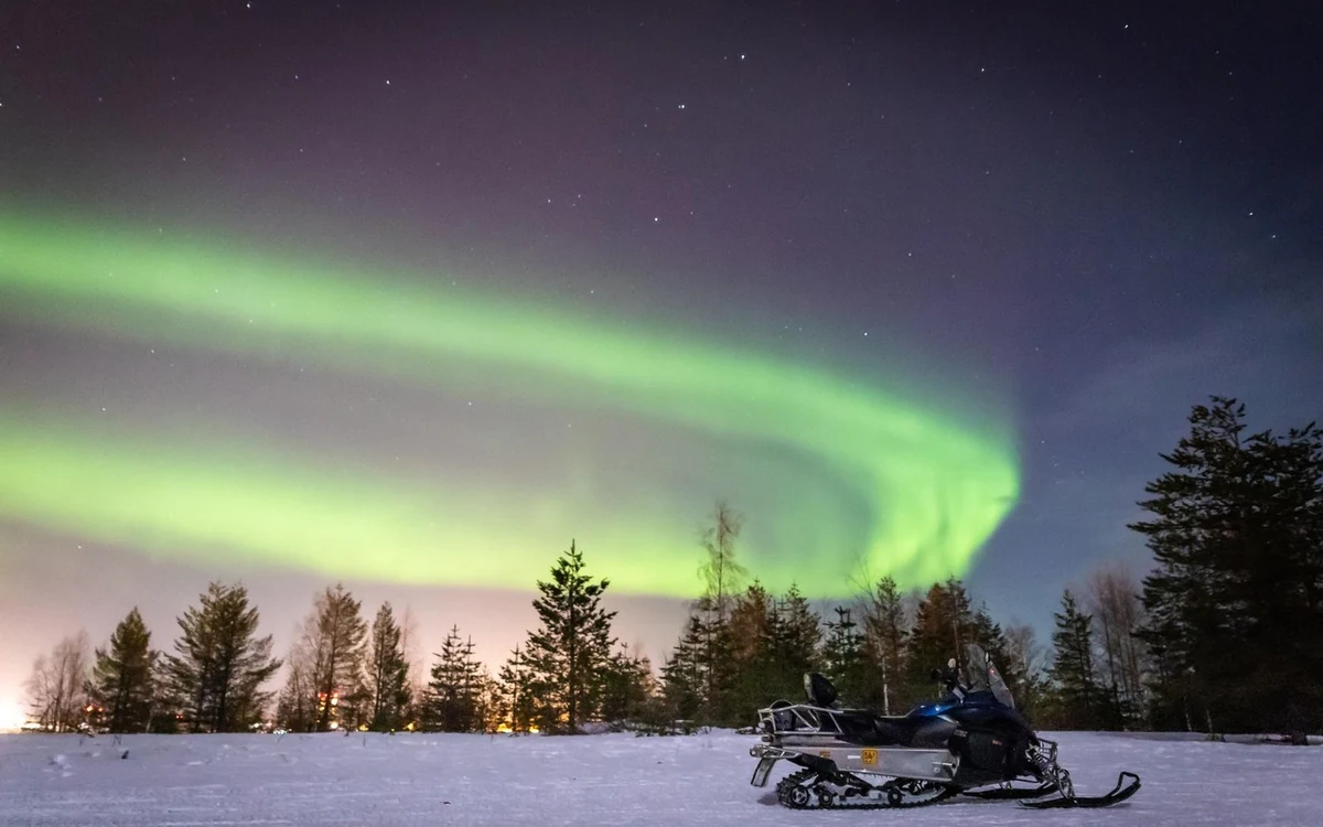 A snowmobile parked on open snow with a green aurora band over a line of spruce