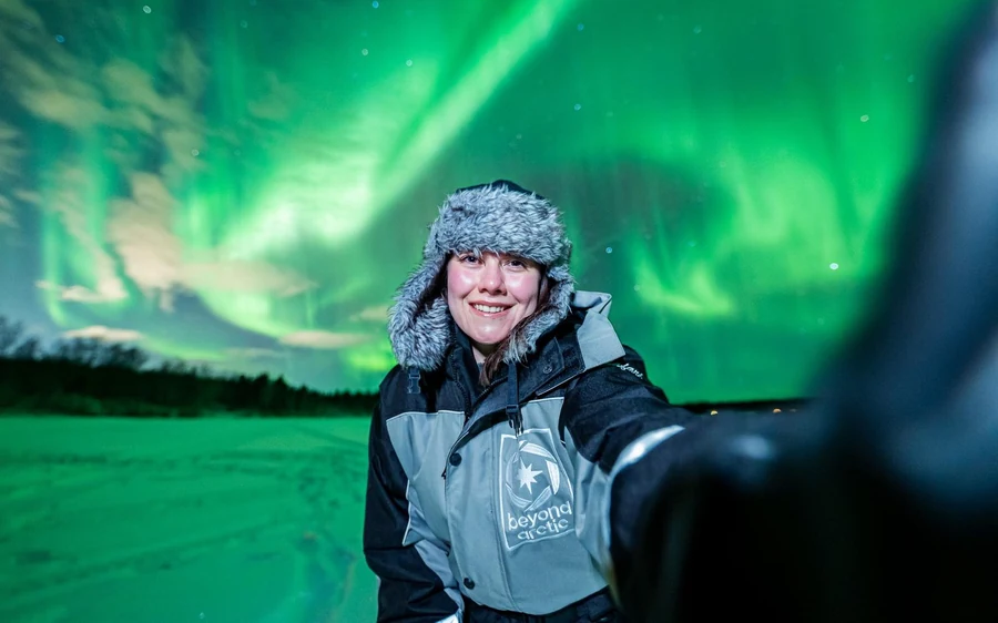 A smiling woman in a fur-lined hat and thermal suit taking a selfie under a bright green sky