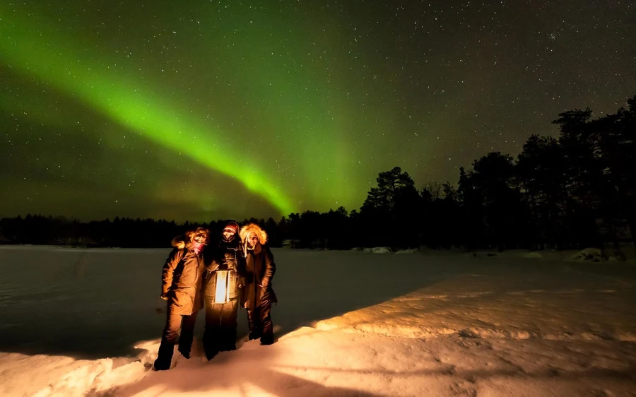 Three people in thick parkas standing on snow with a lantern under a green aurora above the treeline
