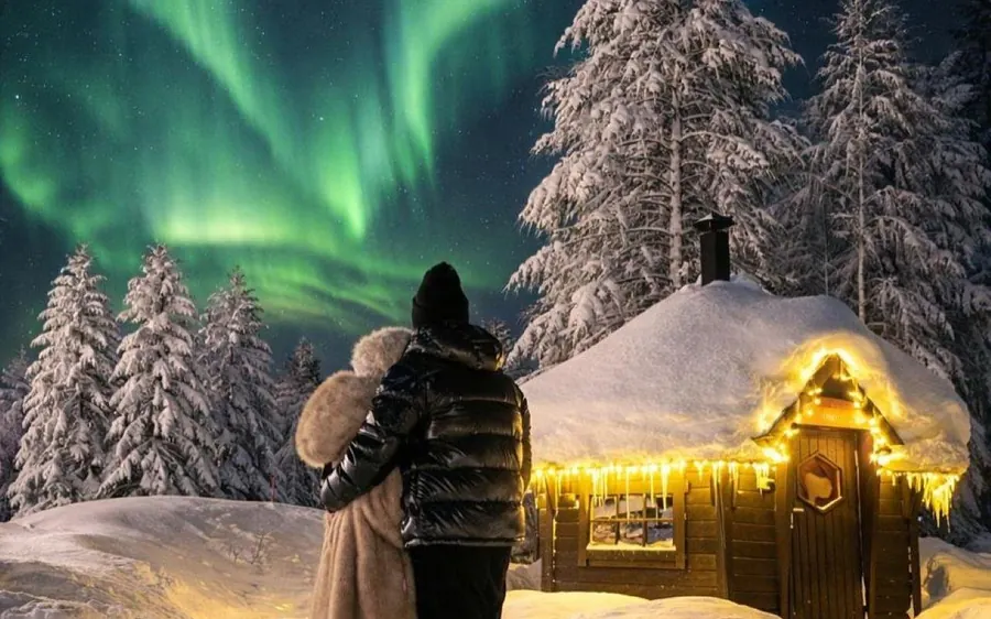 Two people in winter coats by a snow-covered wooden hut strung with warm lights under a green sky