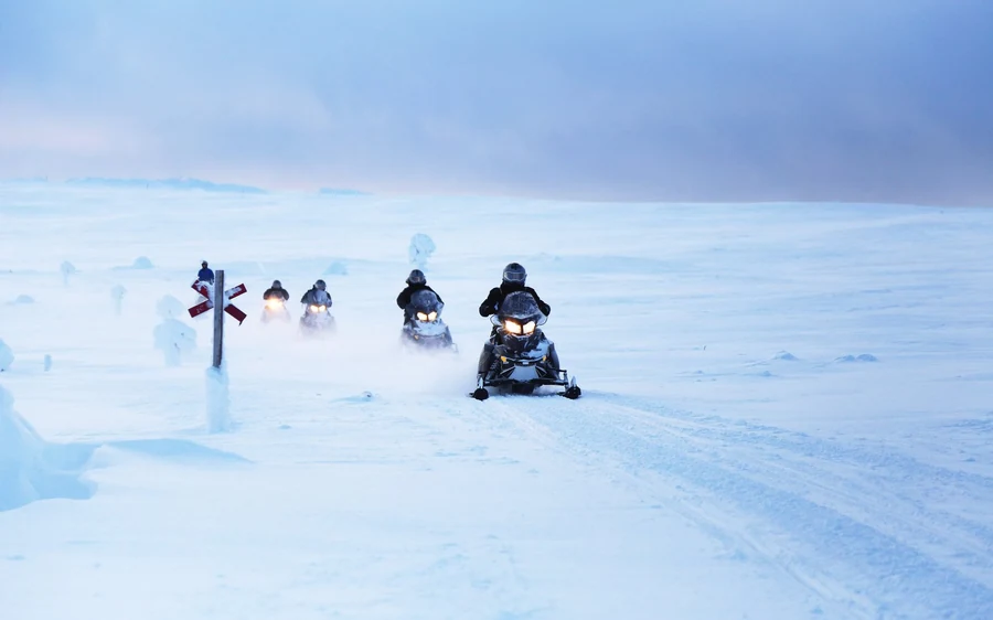 A line of snowmobiles with headlights on crossing open wind-blown snow in dim light